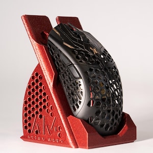 May include: A red 3D-printed mouse stand with a honeycomb pattern and the text "AM ASTRA MODS". A black gaming mouse is placed in the stand. The stand is designed to hold a computer mouse.
