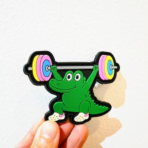 May include: A rubberised patch featuring a green cartoon alligator lifting a barbell overhead. The barbell has pink and blue weight plates on each side. The alligator is wearing white shoes with pink accents and has a black outline border.