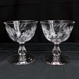 May include: A pair of clear champagne coupe glasses featuring etched leaf and vine patterns. Each glass has a circular base, an ornate stem, and a broad bowl. The etched design is displayed on the bowl of each glass.