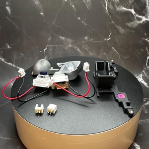 Może przedstawiać: Zbliżenie na rozłożony mechaniczny przełącznik klawiatury. Przełącznik jest wykonany z czarnego i srebrnego plastiku i ma przymocowany do niego czerwony i czarny przewód. Są też dwa białe plastikowe złącza i mały czarny plastikowy element z fioletową naklejką.