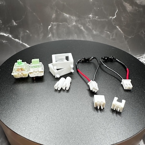 May include: A set of mechanical keyboard switch components including two green and black mechanical keyboard switches, two white plastic switch housings, two white plastic switch stabilizers, and two white plastic connector wires.