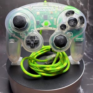 Pode incluir: Um comando Nintendo GameCube transparente com uma placa de circuito interna verde visível, joysticks pretos, um pad direcional preto e botões de ação pretos. Um cabo entrançado de cor verde néon e amarelo está enrolado à frente.