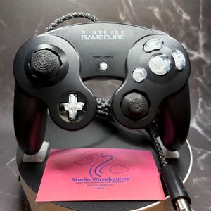 Op de afbeelding: Een zwarte Nintendo GameCube-controller met aangepaste wit gemarmerde knoppen en een D-pad. De controller heeft een zwarte gevlochten kabel en wordt getoond met een roze kaart met de tekst Studio Wavebounce, Custom Gamecube Controllers and Peripherals, EAST VILLAGE, NYC, 2024.