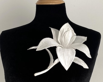 May include: A white lily flower brooch with a textured, shimmering surface and pointed petals. The flower features a central stamen and a curved stem extending to the left, pinned onto a black velvet-textured display form with a wooden neck.