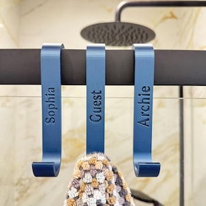 May include: Three blue shower hooks with the names "Sophia", "Guest", and "Archie" engraved on them. The hooks are hanging on a black shower rod.
