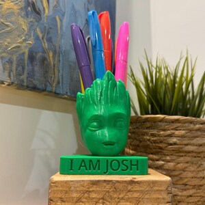 May include: A green 3D printed Groot head pencil holder with five colourful pens sticking out of the top. The holder is sitting on a wooden base with a green plaque that reads "I AM JOSH".