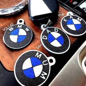 May include: Close-up of several BMW-themed keychains. The keychains are round with a black textured border and a white and blue logo. Some keychains have the text "MUM", "DAD", and "KAM". A car key and other car accessories are also visible.