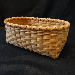May include: A woven basket made of natural materials, with a light brown color and a rectangular shape. The basket has a woven rim and is empty.