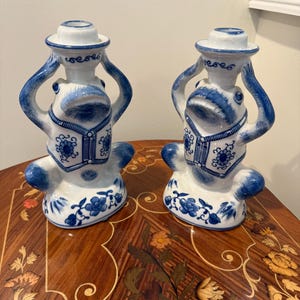 May include: Two ceramic candle holders in a whimsical frog design. The candle holders are white with blue accents, featuring a detailed floral pattern. The frog-shaped figures have their arms raised, holding the candle cup.