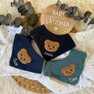 Könnte beinhalten: Drei Baby-Lätzchen in Marineblau, Türkis und Blau, jedes mit einem Teddybär-Applikation. Die Lätzchen sind mit den Namen Theo, Amelie und Lina personalisiert. Im Hintergrund ein Holzschild mit den Worten "BABY LÄTZCHEN".