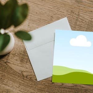 May include: A greeting card with a landscape design featuring a blue sky, white cloud, and green rolling hills. The card is next to a white envelope on a wooden surface, with a green plant in the background.