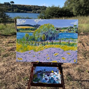 Può includere: Dipinto paesaggistico impressionista su cavalletto. L'opera raffigura una scena vibrante con un lago blu, alberi verdi e campi gialli sotto un cielo blu. Il dipinto è esposto all'aperto su un cavalletto di legno.