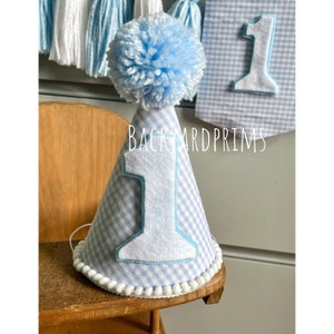 May include: A blue and white gingham party hat with a white felt number one and a blue pom pom on top. The hat is sitting on a wooden surface. "Backyard Prims" is written on the banner behind the hat.