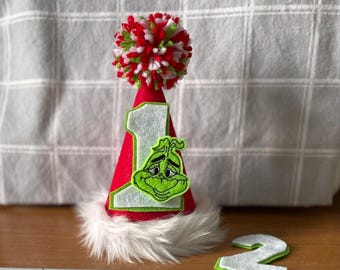 May include: A red cone-shaped party hat featuring a white felt number one, a green embroidered character face, and a white faux fur trim at the base. The top is decorated with a red, white, and green pom-pom. A separate white felt number two is nearby.