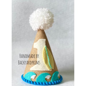 May include: A burlap-textured birthday party hat with a white pom-pom on top. The hat features a felt number 1, a surfboard, and ocean wave accents. The text "HANDMADE BY BACKYARDPRIMS" is printed on the hat.