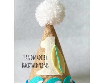May include: A burlap-textured birthday party hat with a white pom-pom on top. The hat features a felt number 1, a surfboard, and ocean wave accents. The text "HANDMADE BY BACKYARDPRIMS" is printed on the hat.