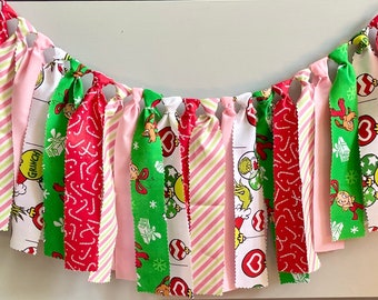 May include: A festive fabric banner featuring alternating strips of pink, red, and green fabric. The banner includes patterns of candy canes, stripes, and Dr. Seuss's Grinch characters, perfect for holiday decorations. The banner is tied together with pink string.