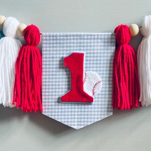 May include: A blue and white gingham banner with a red felt number one and a red felt baseball. The banner is decorated with blue, white, and red yarn tassels.