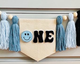 May include: A blue and black tassel garland with a white banner that says "ONE" with a blue smiley face.