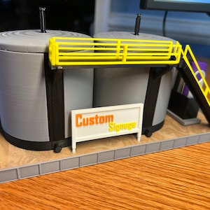 May include: A 3D printed model of a small industrial facility with two gray tanks, a yellow railing, a yellow ladder, and a white sign that reads "Custom Signage".