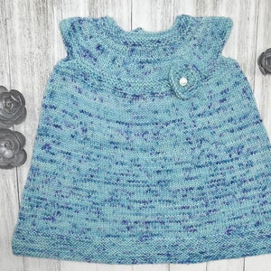Könnte beinhalten: Ein hellblaues gestricktes Babykleid mit einer weißen Blumenverzierung. Das Kleid hat einen gewellten Ausschnitt und ist ärmellos.