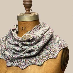 Peut inclure: Un col tricoté à la main dans des tons de gris, de violet et de vert, présenté sur un mannequin en bois. Le col présente un bord festonné et un motif en tricot ajouré texturé. Le mannequin a une base en bois et un dessus en métal.