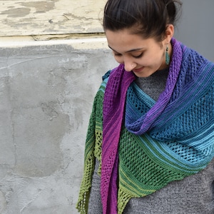 以下が含まれることがあります： グレーのセーターを着た女性が、レース模様のカラフルなクロシェのショールを巻いています。ショールは、パープル、グリーン、ブルーのグラデーションになっています。