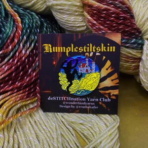 May include: A black and yellow enamel pin featuring a silhouette of a figure standing on a cliff with a castle in the background. The pin is round and has a blue border. The text "Rumplestiltskin" is printed in black on a brown background. The text "deSTITCHnation Yarn Club" is printed in black on a white background. The text "@wonderlandyarns" is printed in black on a white background. The text "Design by @rrutshaffr" is printed in black on a white background.