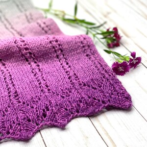 May include: A purple and gray ombre knitted scarf with a lacy pattern. The scarf is folded and lying on a white wooden surface with pink flowers.