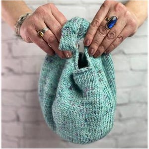 May include: A hand-knitted, drawstring bag in a speckled aqua color. The bag is held open, showcasing its interior. The yarn has flecks of purple and blue. The bag's handle is also knitted.