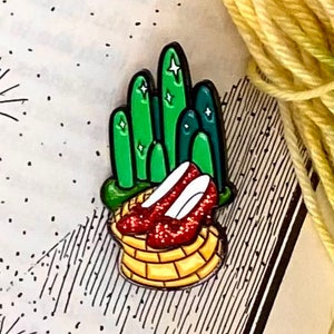 Peut inclure: Une épingle représentant une chaussure rouge en rubis avec une route de briques jaunes et une ville d'émeraude verte en arrière-plan.