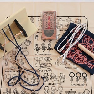 Puede incluir: Una estera de tela beige con una variedad de nudos atados con cuerda azul y blanca. La estera tiene ilustraciones de diferentes nudos y sus nombres, incluyendo "Slippery Hitch", "Clove Hitch", "Anchor Bend", "Loop Knot" y "Pile Hitch".