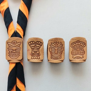 Op de afbeelding: Vier rechthoekige houten badges met gegraveerde tiki-gezichten. De badges zijn bevestigd aan een zwart-oranje gestreepte das.