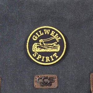 Może przedstawiać: Szara, płócienna torba listonoszka z brązowym paskiem skórzanym i klamrami. Torba ma naszywkę z żółtym haftem z napisem "Gilwell Spirit" i grafiką przedstawiającą kłodę i siekierę.
