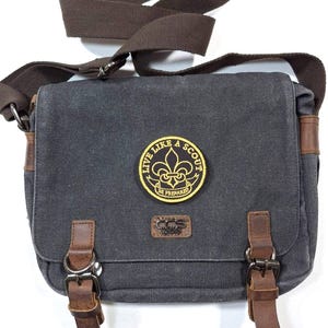 May include: A gray canvas messenger bag with brown leather straps and buckles. The bag has a patch with the text "Live Like A Scout" and a fleur-de-lis design. The text "Be Prepared" is also on the patch.