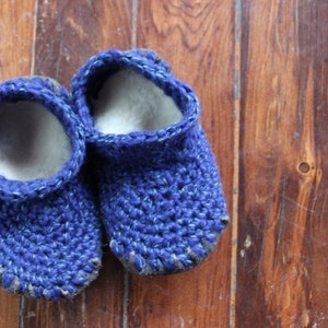 May include: A pair of blue crocheted slippers with a white lining. The slippers are on a wooden floor.