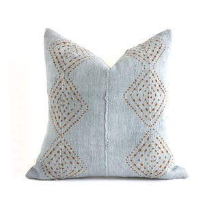 May include: A square, light blue decorative pillow with a geometric diamond pattern in a warm brown color. The pillow has a textured appearance and is likely made of fabric. The design is symmetrical, with the diamond pattern repeated on both sides.