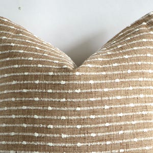 May include: A decorative pillow with a brown woven fabric and horizontal white dotted stripes. The pillow has a textured appearance and a neutral color palette, suitable for various home decor styles.