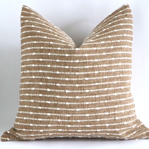 May include: Square decorative pillow with a tan and white striped pattern. The pillow has a textured appearance with horizontal stripes of tan fabric and evenly spaced white dots. The pillow is a neutral color and would complement any home decor.