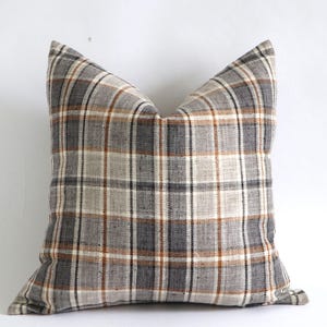 May include: A square pillow with a plaid pattern in shades of gray, brown, and cream. The pillow has a soft, textured appearance and a classic, neutral design. The pillow is a decorative home accent.