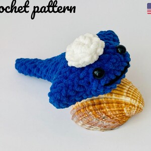 May include: Crochet pattern for a blue and white amigurumi whale with black eyes. The whale is sitting on a seashell.