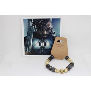Könnte beinhalten: Schwarzes und goldenes Perlenarmband mit einem Totenkopf-Anhänger. Das Armband ist handgefertigt mit Liebe. Das Bild zeigt auch ein Foto eines Mannes in einem schwarzen Anzug.
