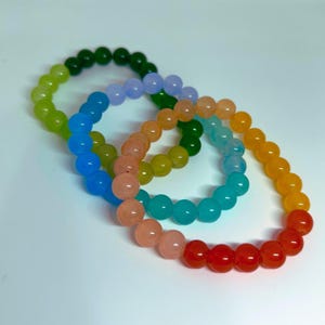 May include: A collection of beaded bracelets in a rainbow of colors. The bracelets feature a gradient of colors, including green, blue, orange, and red. The beads are round and appear to be made of a smooth material.