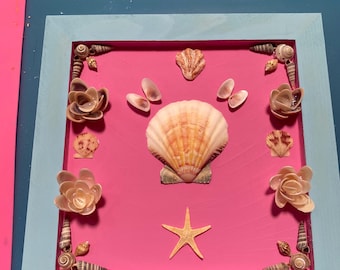 May include: A framed seashell art piece with a pink background. The frame is light blue with a white inner border. The seashells are arranged in a symmetrical pattern with a large scallop shell in the center. There is a starfish below the scallop shell.