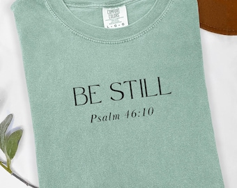 May include: A sage green t-shirt with the words "BE STILL" and "Psalm 46:10" printed in black. The shirt is folded and has a tag that says "Comfort Colors". A brown hat is in the background.