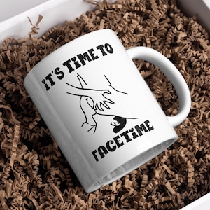 Peut inclure: Une tasse en céramique blanche avec un graphique noir de deux mains tenant un téléphone et le texte "It's Time To FaceTime".
