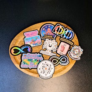 May include: A collection of colorful vinyl stickers featuring various designs including a rocket, an octopus, a light switch, and infinity symbols. The stickers display text such as Hyperfocus Activated, Chaos Coordinator, and I'm not procrastinating.