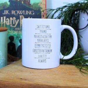 Könnte beinhalten: Eine weiße Keramiktasse mit schwarzem Text, der "SWT.T.CT.BMO YYH.M.D. YKHAST.HCOAT.PM YBRHLAPFS D.T.M.N.T.YG.T.CF.T.B EXPECTO PATRONUM LA.A.T.T? ALWAYS" lautet.