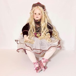 May include: A porcelain doll with long blonde curly hair, wearing a burgundy and gold dress, a white tulle skirt, and pink ballet flats. The doll has a burgundy hat and is seated with legs crossed.