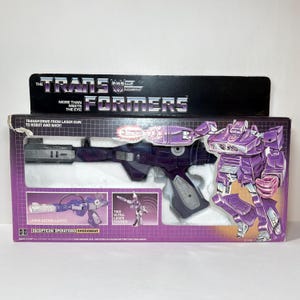 May include: A purple and grey Transformers Shockwave toy in its original packaging. The box features the Transformers logo, the text More Than Meets The Eye, and illustrations of the toy in both laser gun and robot forms with laser action lights.